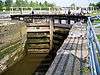 A canal lock with sandstone walls and wooden gates seen from its midpoint; the gates are closed and water is held at a higher level beyond them; railings run along the top of the gates and on the wall to the right