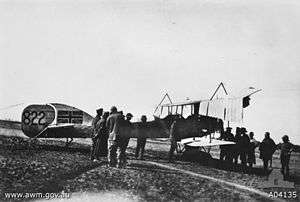 A group of men gather around a biplane. The Union Jack has been painted on to the tail of the aircraft.