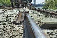 A view along the top surface of a rail with another piece of rail twisted around it a short distance away and a freight car on the track behind it. The ties and ballast in the foreground are a much lighter color than those elsewhere in the image