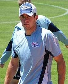 An Australian cricketer wearing blue jersey. An another person can be seen in background.