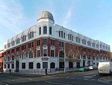 A four-storey building on a corner site, with white stonework on the bottom and top storeys and red brickwork in between. At the corner is a metal dome on an octagonal base.