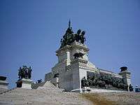 Photograph of a white stone steps leading up to a large, altar-like monument in white marble with bronze sculptural decorations that include bronze braziers at the corners, a bronze frieze in high relief at the base and bronze figures surrounding a chariot on a high, white marble plinth in the center