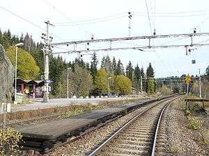 A small station with two platforms; above are three overhead wires and the area is surrounded by forest