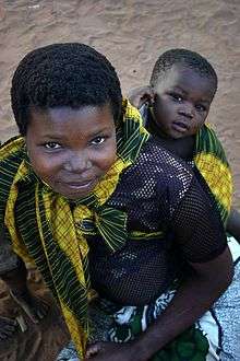 A young mother smiles up at the camera. On her back is her baby gazing at the camera with an expression of lively interest.