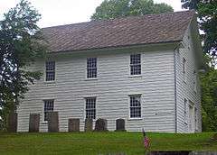 A two-story white building with a pointed roof, seen from the side, with gravestones in front, similarly austere