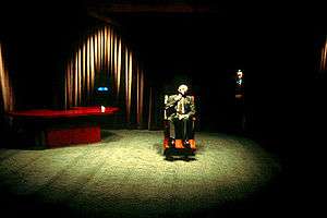 A short, strange-looking man seated in a large wooden wheelchair under an intense beam of light in a large and sparsely furnished room; a desk is in a far corner and the walls are covered in curtains