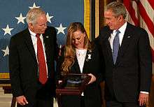 A color image of Murphy's parents standing next to President George Bush in front of an American flag. They are holding Murphy's Medal of Honor in a display case and are looking down at it.