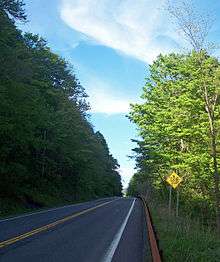 A two-lane road in a wooded area where one side of the road has a steep slope. A "Fallen Rock Zone" sign is visible on the right of the road