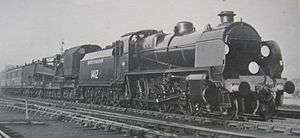 A side-and-front view of a 2-6-0 steam locomotive coupled to a breakdown crane in a yard. The locomotive features smoke deflector plates either side of the boiler and there are white route discs on the front.