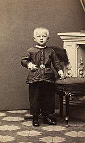 an unsmiling fair-haired child stands upright, his left hand resting on a stool, in front of an ornate fireplace.