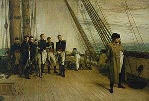 A group of men in military uniform and formal clothes stand to the left, looking towards a single man in a greatcoat and bicorne hat stood by the rail of a ship looking out to sea