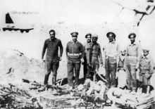 Eight men in dressed in military fatigues standing before an organized assembly of weapons, mostly rifles and mortar. The first man from the left is not wearing a hat, while the remaining seven are wearing hats.