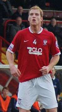 A man with blonde hair who is wearing a red top, white shorts and red socks. He is standing on a grass field.