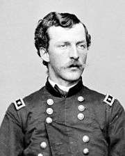 Head and shoulders of a young white man with wavy hair and a mustache, wearing a double-breasted military jacket with a rectangular patch over each shoulder.