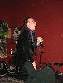 Photograph of a man in a purple shirt and dark suit, holding a microphone.