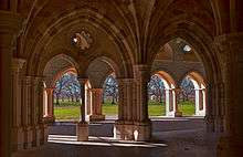 Multiple interior and exterior stone arches frame a view looking out from a chapter house, revealing a view of orchard trees beyond an expanse of bare ground.