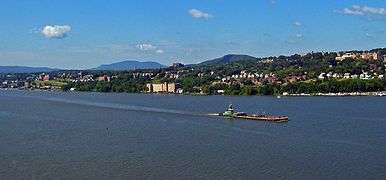 A city on a slope over a body of water with larger buildings at the left and center right. There are low hills in the distance