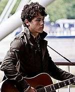 A three-quarter portrait of a male teen looking forward. He has dark, curly hair, wears a black leather jacket, and strums a guitar hung over his shoulder.