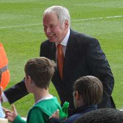 A middle-aged grey-haired man in a dark business suit smiles and talks to people out of frame