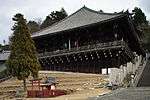 Large wooden building with a pyramid shaped roof. Built on a hill slope with part of the building and veranda supported by poles.