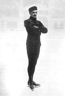 A male figure skater poses with crossed arms for a shot at an indoor ice rink; the background is very blurred.