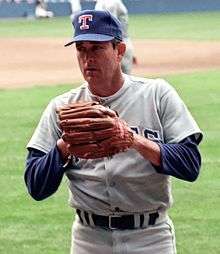 A man in a gray baseball jersey wearing a dark blue baseball cap with a "T" at the centre and a brown Rawlings on his left hand prepares to pitch with the windup.