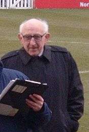 A man with white hair is wearing a black jacket. He is standing on a grass field.