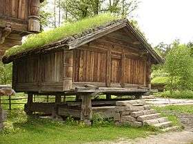 Crude log and timber open-air cabin with a pitched roof on a foundation of rocks, with the second storey overhanging the first.