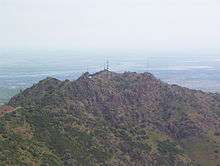 Behind the rocky form of a hill in the foreground, is visible a sizable plain partially obscured by fog and crossed by several winding rivers.