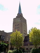 A tall light-coloured stone square tower with a small metal spire; to the left, a smaller building in the same stone with a dark tiled roof