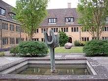 A small pool surrounded by paving slabs; a metal abstract sculpture stands in the pool, with a long post topped by a ribbed "U" shape in front of a segment of a sphere; behind the pool, a lawn with trees and bushes, and two storey buildings with regular windows and dark tiled roofs