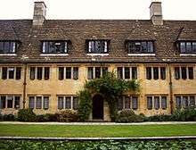 In the foreground, part of the long pool and lawn; behind, a section of a two storey building in light stone, topped with a dark tiled roof and two chimneys; there are dormer windows in the roof; the windows at ground and first floor level are regular and frequent; an open archway surrounded by vegetation in the centre