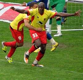 Two men wearing yellow shirts, red shorts and red socks, celebrating on a grass field