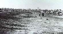 In the distance a group of people walk down a dirt road away from the village. The foreground shows vacant ground.