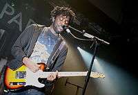 A male vocalist plays electric guitar in front of a mic stand. The stage is lit by white lights,  and its background is mostly black.