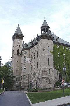 Relatively tall and narrow five storied building of grey stone with a dark roof and an attached clock tower.