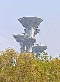 A narrow tower with several round, wider flat-roofed tops seen against a pale blue sky above a group of trees in early spring