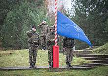 Three soldiers in camouflage stand in salute while a forth raises a blue and white flag on a red and white stripped flagpole.