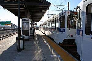 A white train is parked at an island platformed station with canopies and signage visible.