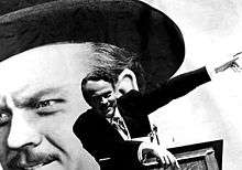 A black-and-white image of a man wearing a suit standing at a podium. His right arm is resting on the podium and is left is pointing up to his left. A large headshot of the same man is visible in the background.