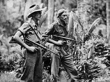 Two men wearing military style uniforms, one wearing a slouch hat, the other a general service cap, carrying weapons in a jungle setting