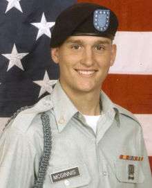 Head and shoulders of a smiling young man in circa 2000 U.S. Army uniform with beret, before a large American flag.