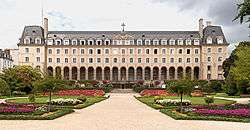 A large four-storey sand-coloured building with 14 arches at the ground-floor entrance and a large landscaped formal garden in front.  The photograph was taken during the summer, so many flowers are in bloom and there are leaves on the bushes and trees.