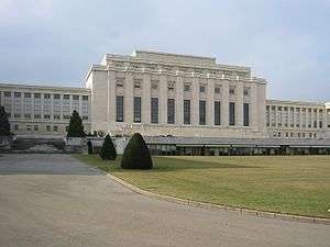 A drive leads past a manicured lawn to large white rectangular building with a columns on it facade. Two wings of the building are set back from the middle section.