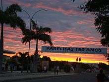 A street in Parelhas, with palm trees and buildings, and a banner stating "150 Anos" (150 years) across the street, at sunset