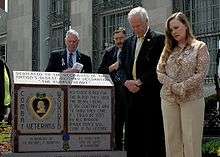 A color picture of Daniel and Maureen Murphy standing next to a monument in front of the Lieutenant Michael P. Murphy Post Office in Patchogue, New York. The monument has a purple heart and some wording inscribed on it and there are two men in the background.