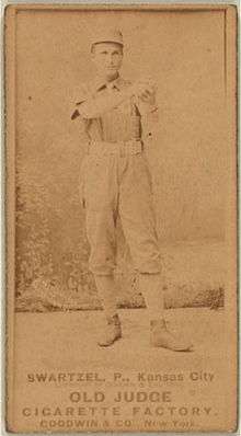 A baseball player is standing in his uniform, holding a baseball.