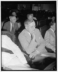 A man in his late forties with graying hair and glasses sits in a chair with his chin rested on his right hand with his right index finger extended. He is wearing a gray jacket and pants with a white shirt and patterned tie