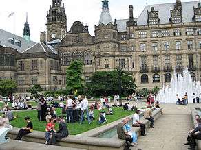 View across a garden containing people enjoying a sunny day towards a large Victorian building with a clock tower