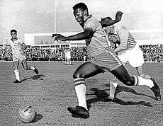 A dark-skinned young man in soccer attire is pictured in mid-stride as he sprints with the ball past an opposing player, who looks tired and has given up attempting to chase him.